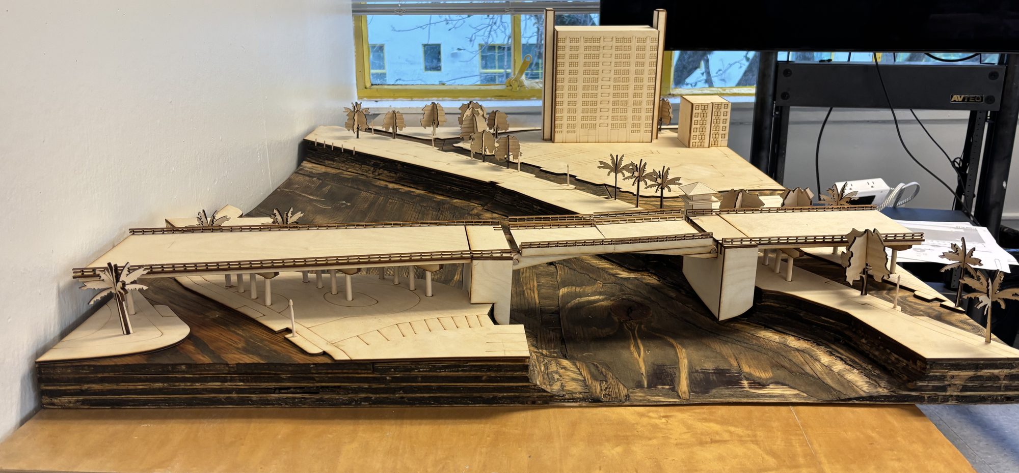 Laser-cut wood site model of a river crossing: a bridge deck on piers over a stained river, a tower and palms on the bank